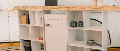 A white minibar with a wooden top, a small refrigerator, and shelves with items like mugs and a trash bin.