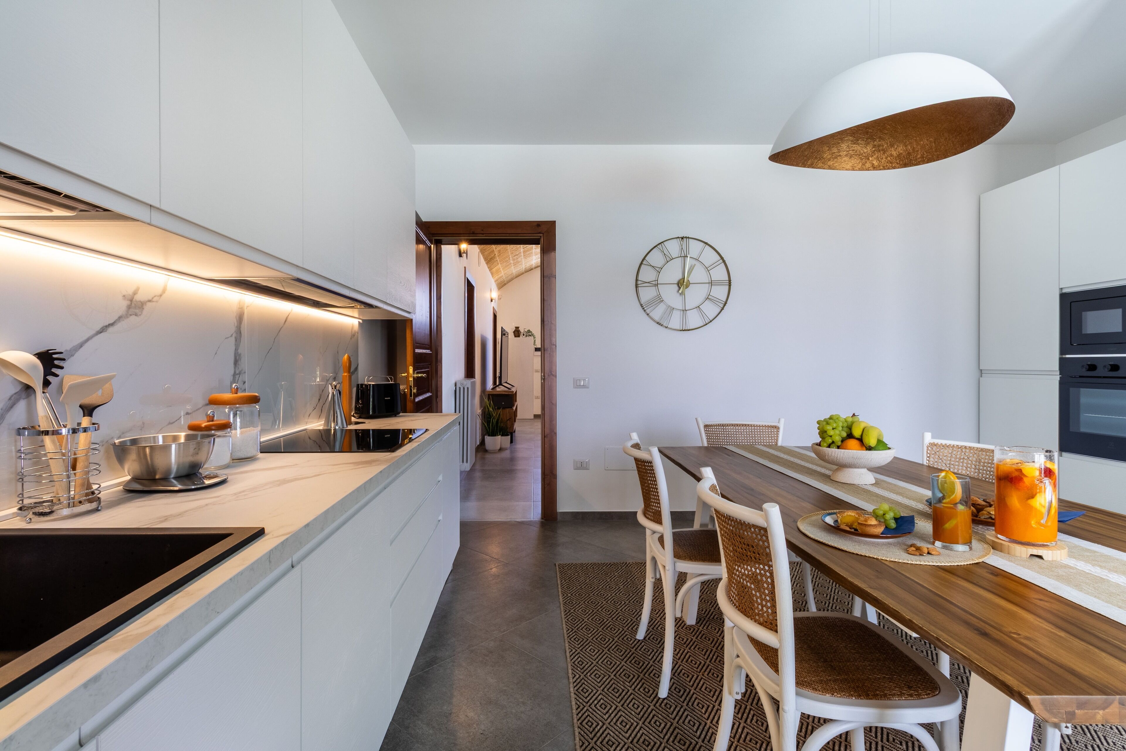 A modern kitchen with white cabinets, a wooden dining table, and a large clock on the wall.