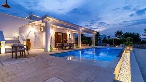 Un área de piscina moderna al aire libre con zona de estar techada, piso de piedra y ambiente bien iluminado.