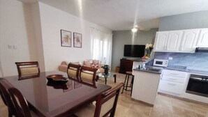 A dining area with a wooden table and chairs, a kitchen with white cabinets, a flat-screen TV, and a small island with a microwave.