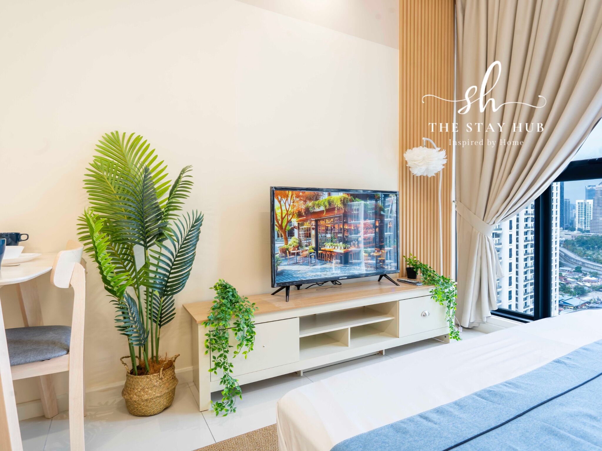 Een moderne hotelkamer met een grote televisie op een houten standaard, een bed met een blauwe spreidek, een witte stoel en een groot raam met uitzicht over de stad.