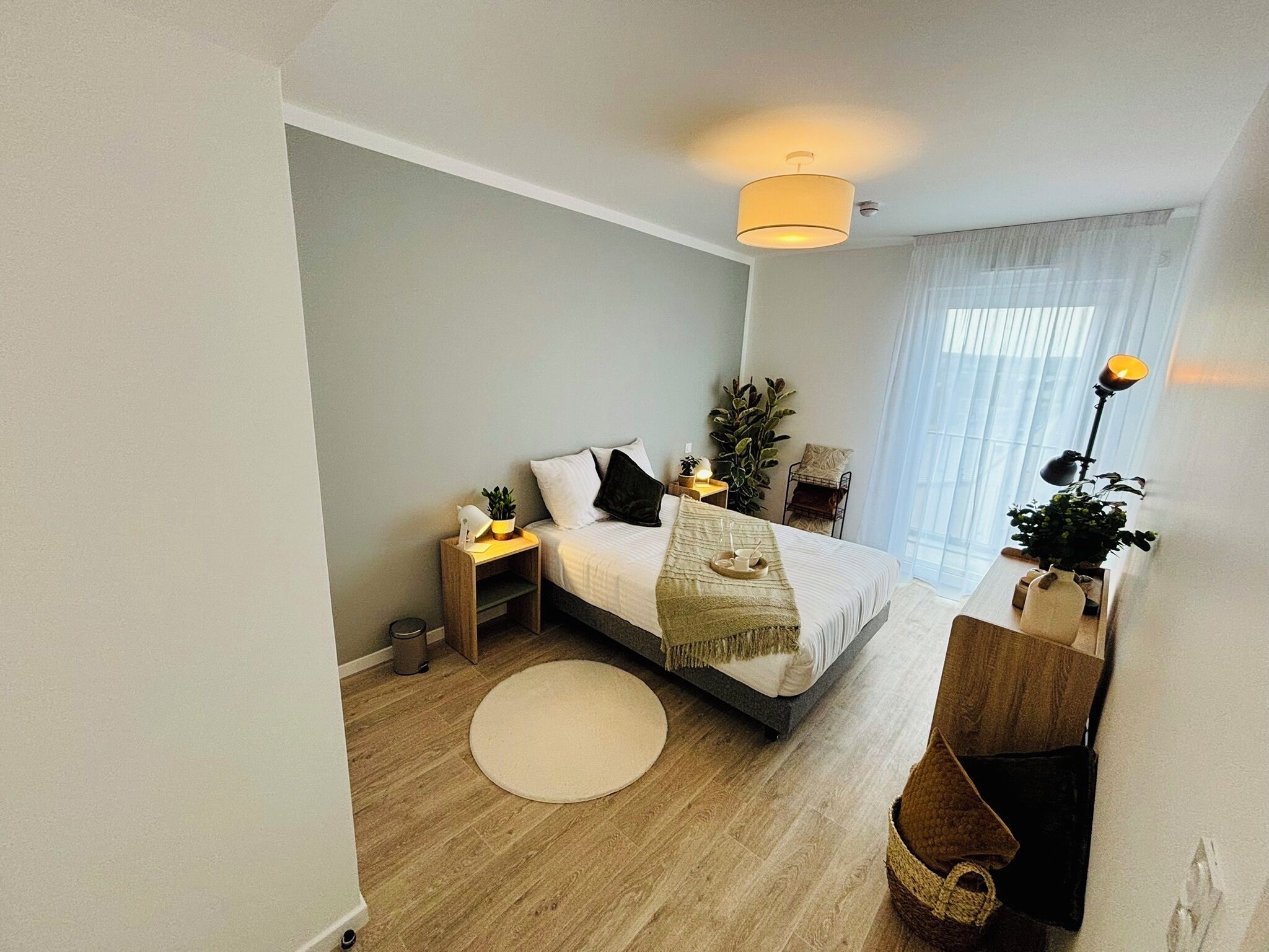 Quarto moderno com cama, mesas de cabeceira, luminária, planta em vaso e janela com cortinas.