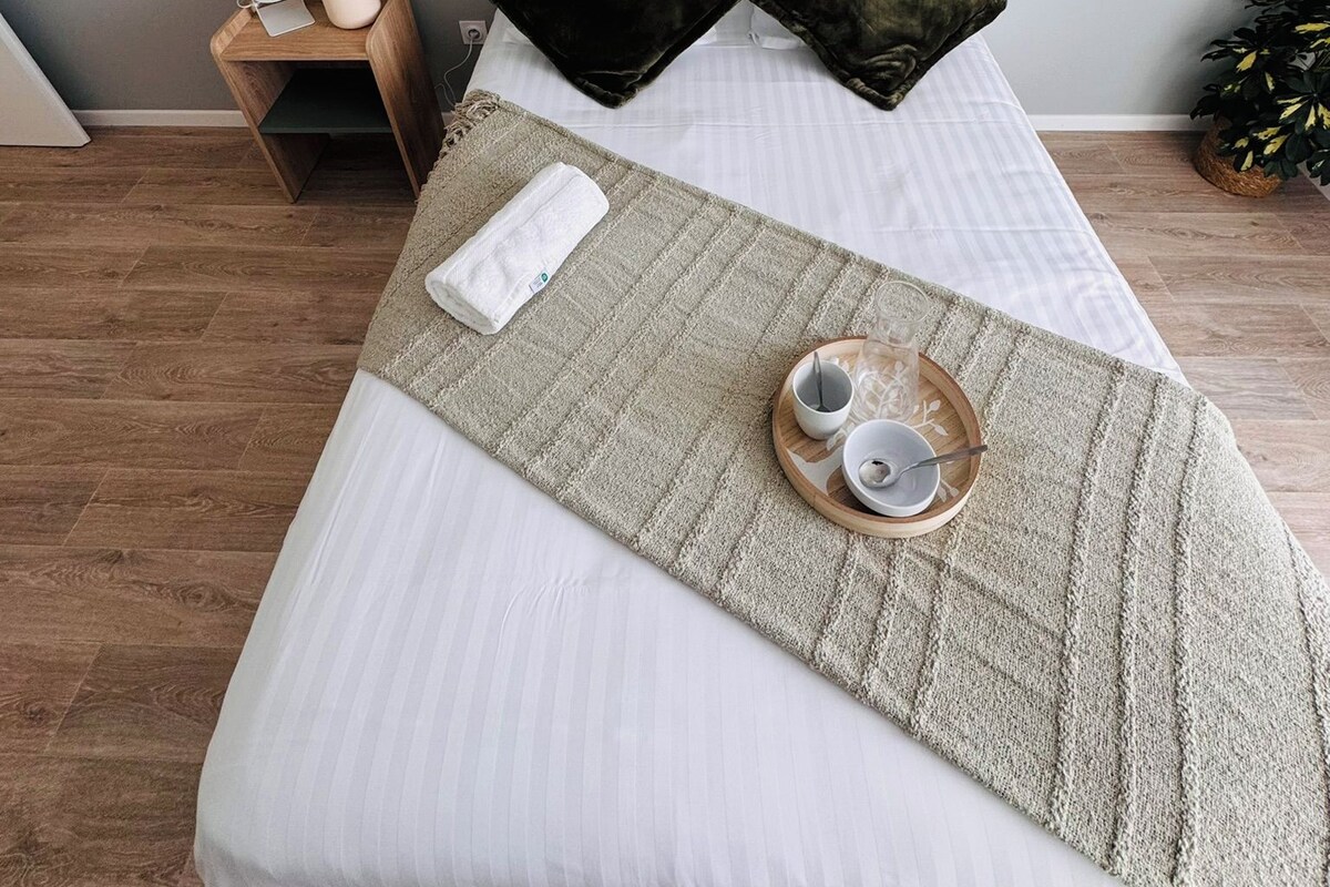 Ein ordentlich bezogenes Bett mit weißer Bettwäsche, zwei dunklen Kissen, einem weißen Handtuch und einem Tablett mit Tasse und Untertasse.