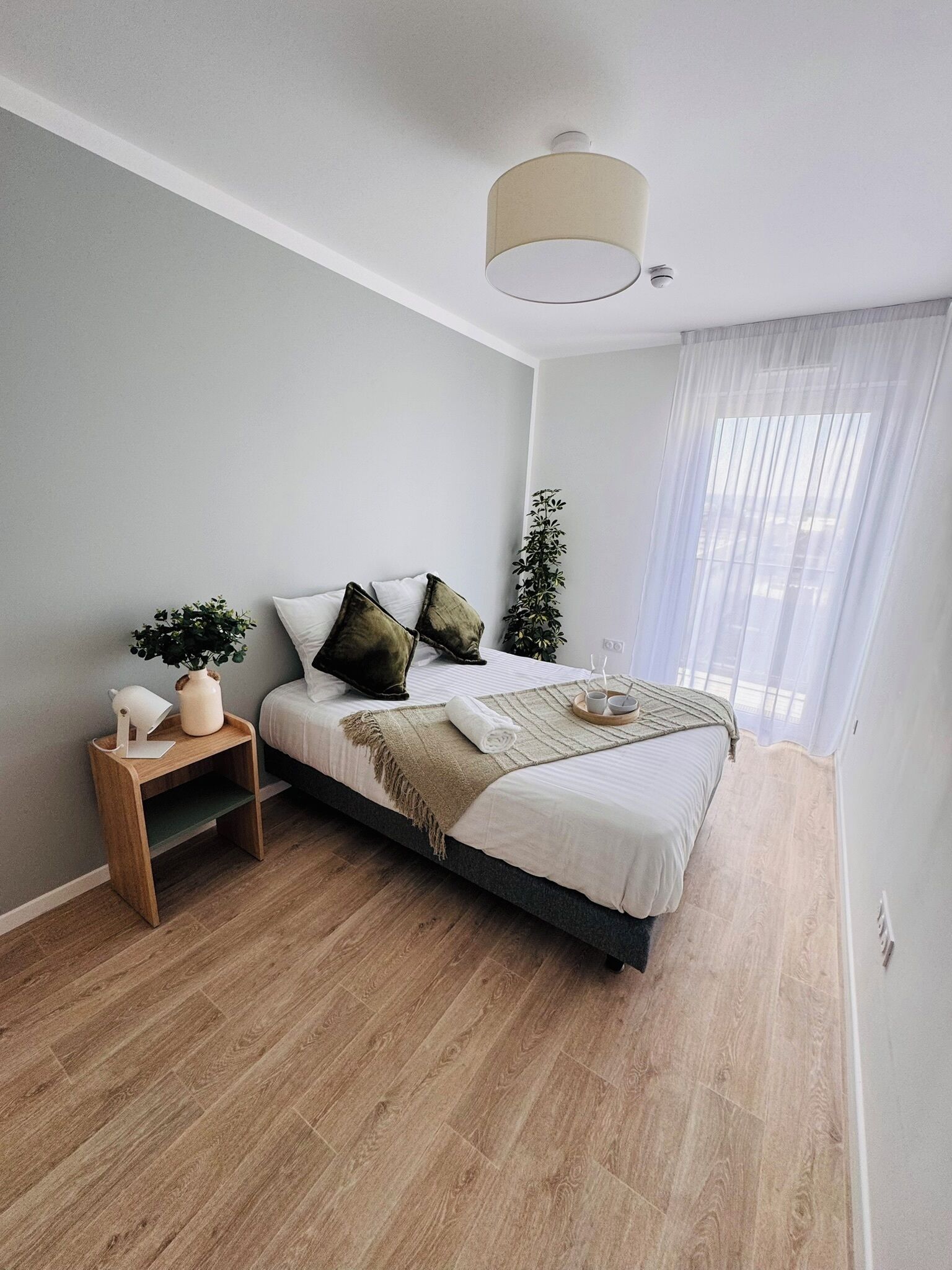 A modern bedroom with a bed, bedside table, and a large window with sheer curtains.