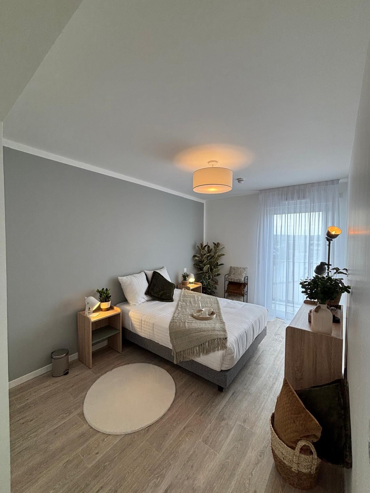 Un dormitorio moderno con cama, mesitas de noche, lámpara de noche, lámpara de pie, una planta en maceta y una ventana con cortinas traslúcidas.