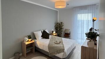 Un dormitorio moderno con cama, mesitas de noche, lámpara de noche, lámpara de pie, una planta en maceta y una ventana con cortinas traslúcidas.
