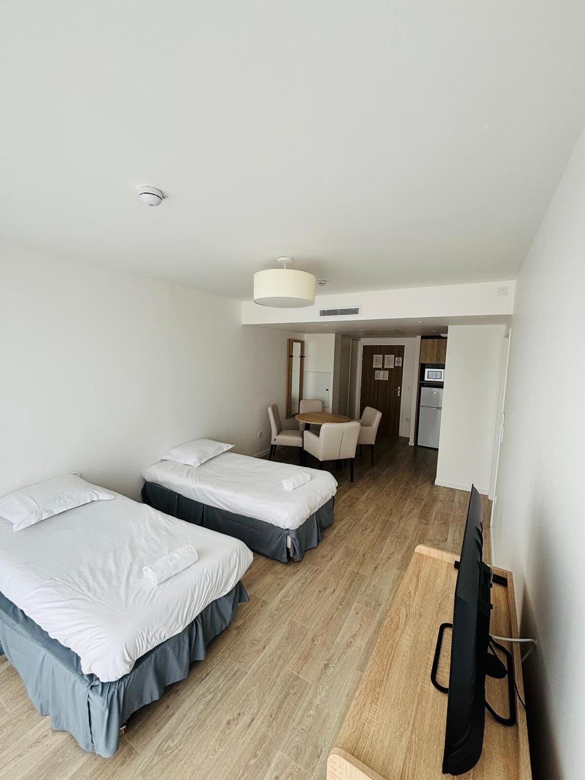 A hotel room with two beds, a desk, a chair, a TV, and a kitchenette.