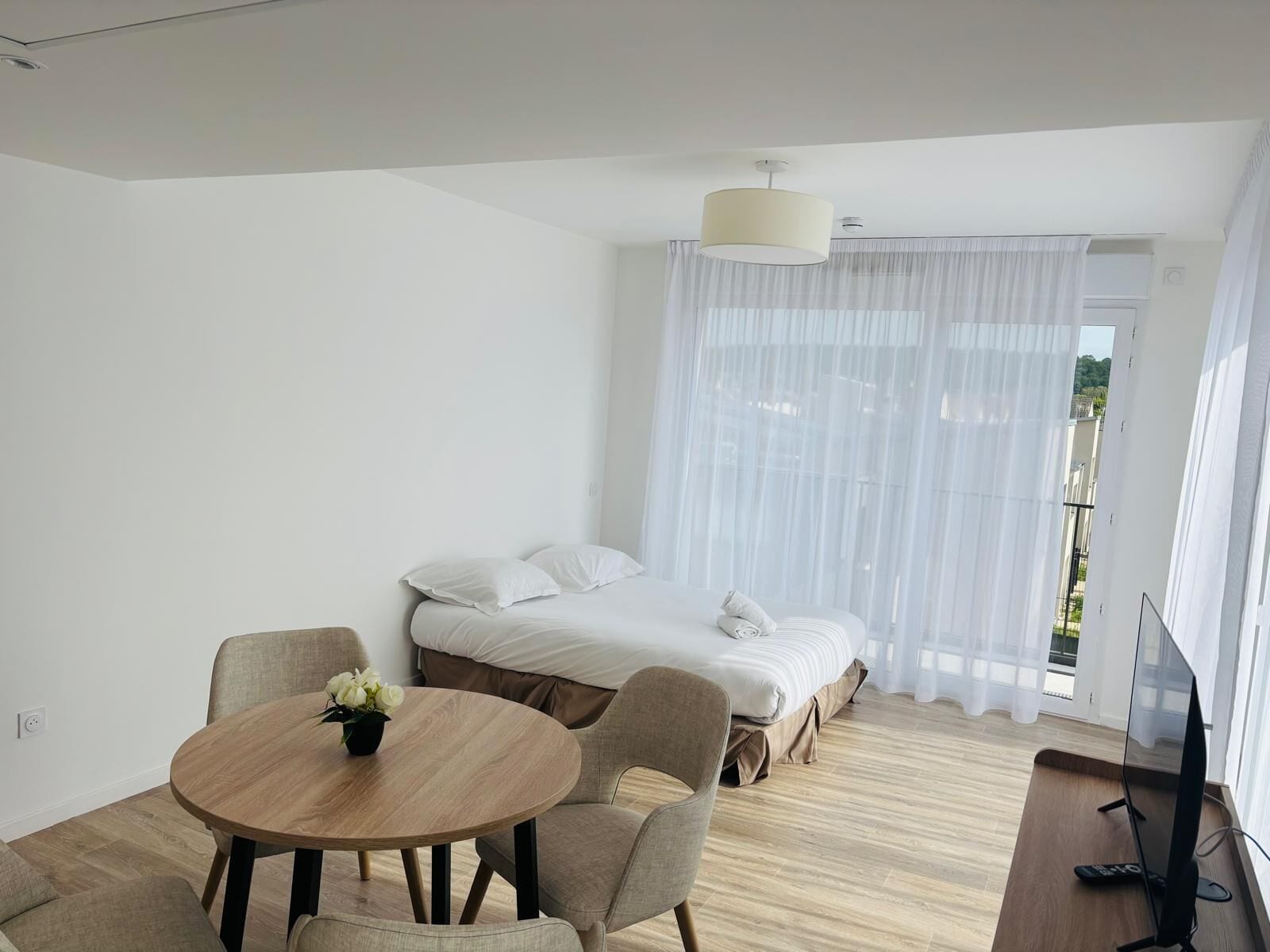 A modern hotel room with a bed, a dining table with chairs, a TV, and a window with sheer curtains.