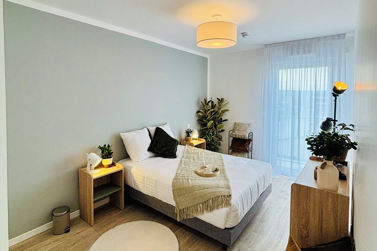 Een moderne slaapkamer met een bed, nachtkastjes, een rond vloerkleed en een groot raam met gordijnen.