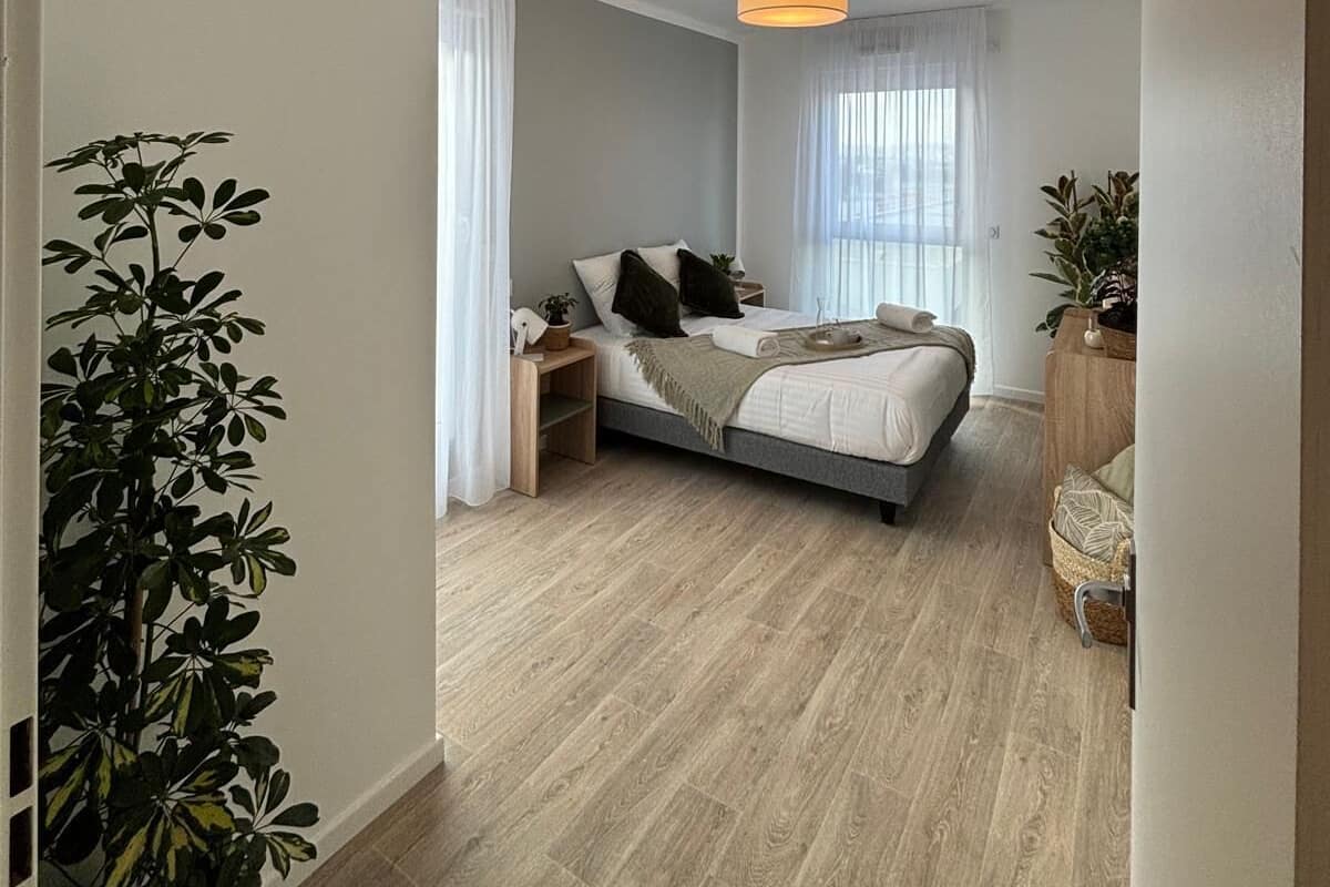 Een slaapkamer met een bed, nachtkastjes, een hanglamp en een raam met dunne gordijnen.