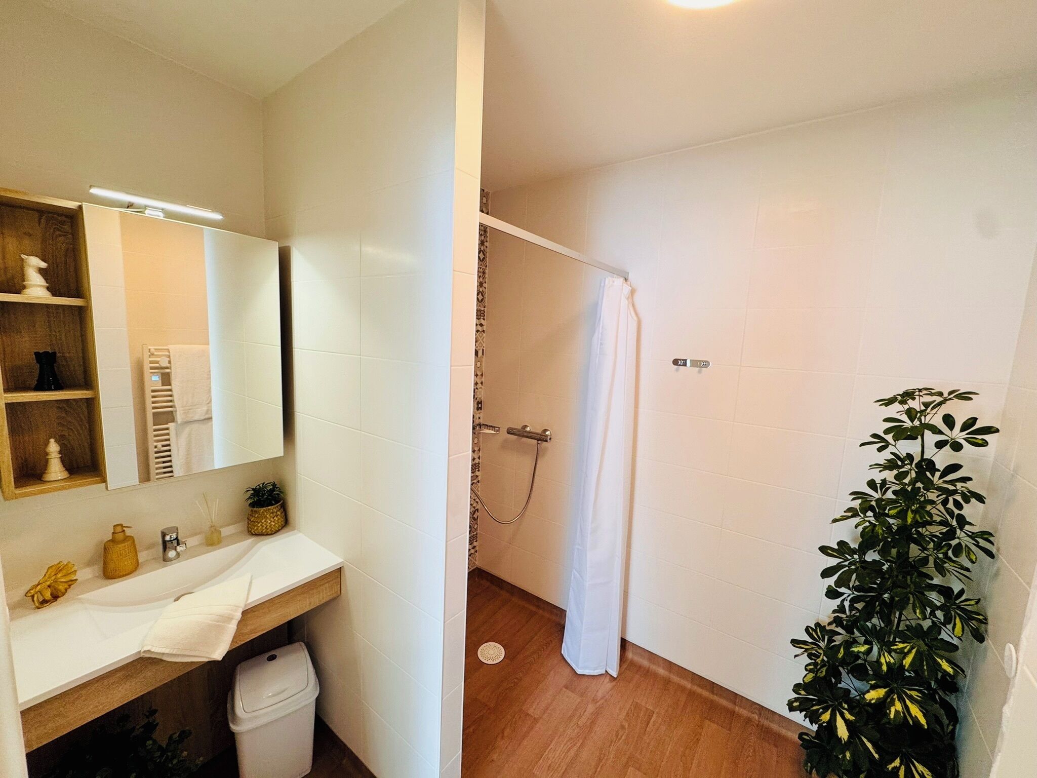 Une salle de bain avec un lavabo blanc, un miroir et un rideau de douche. Il y a une plante sur le côté droit et une poubelle sous le lavabo.