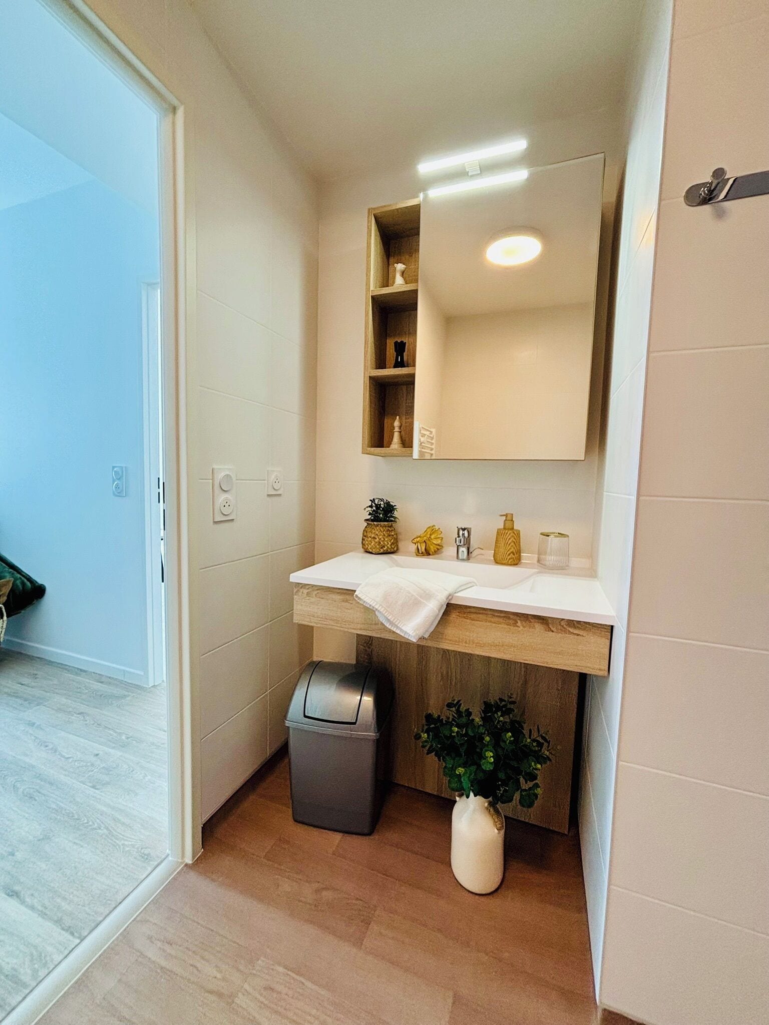 A bathroom with a white sink, wooden vanity, a mirror, a trash bin, and a potted plant.