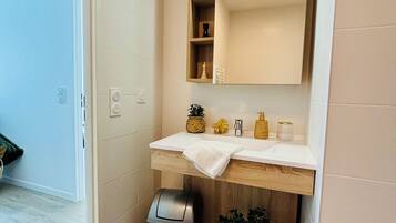 A bathroom with a white sink, wooden vanity, a mirror, a trash bin, and a potted plant.