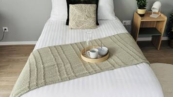 Una cama bien hecha con un edredón blanco, un plaid de punto verde y una bandeja con una taza y un vaso.