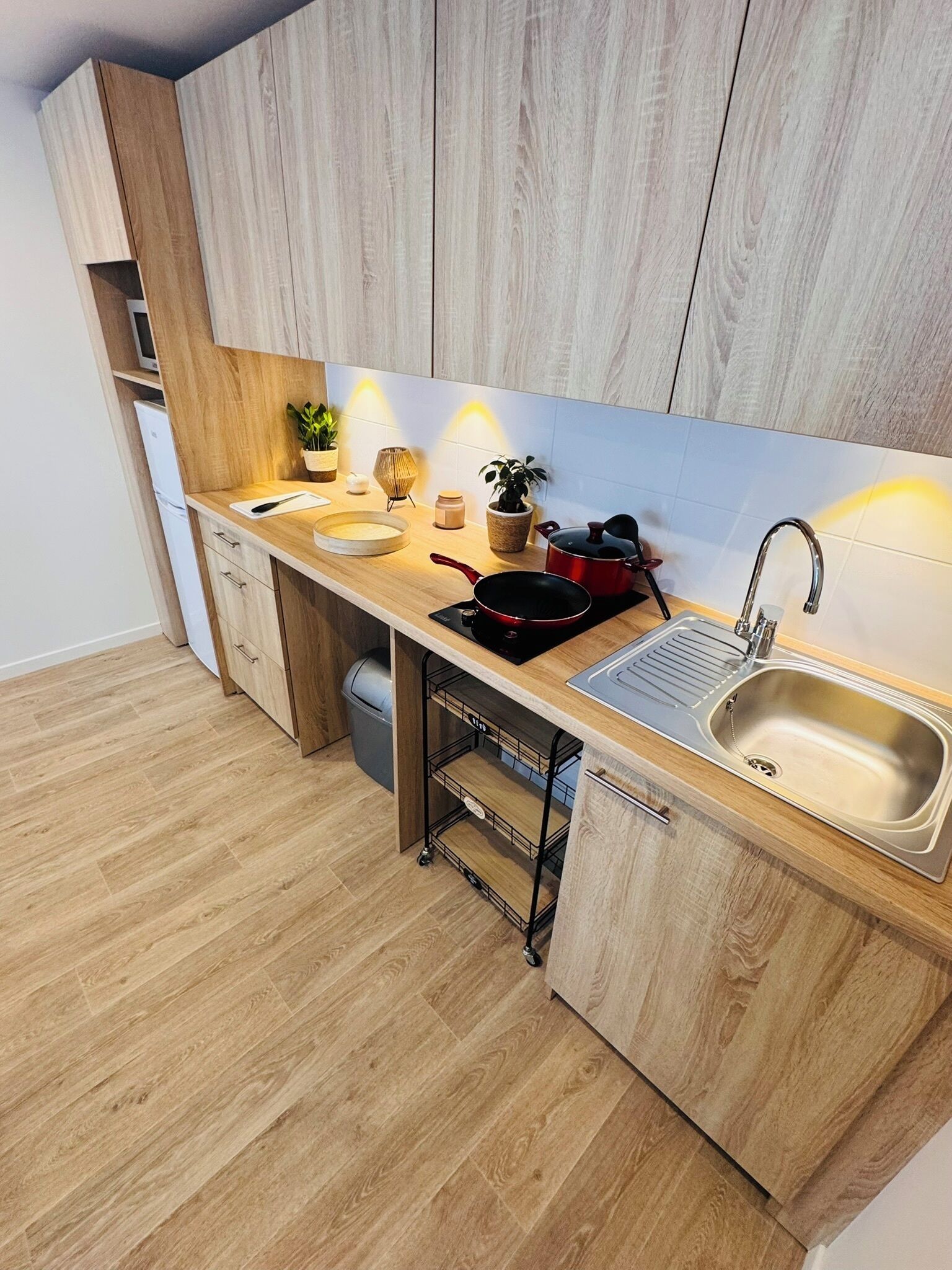 Une cuisine moderne avec des meubles en bois, un évier en acier inoxydable, une poêle rouge et une petite plante en pot.