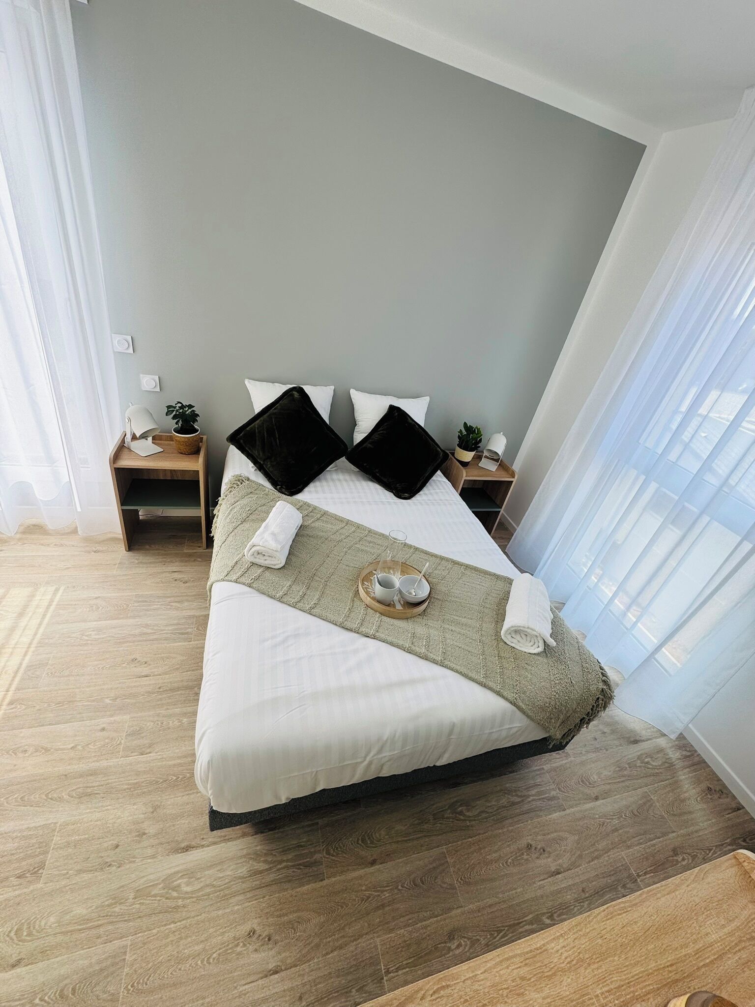 A neatly made bed with a white comforter, two black pillows, and a green throw blanket. There are two bedside tables with lamps and a small potted plant on each. The room has light wooden flooring and light grey walls.