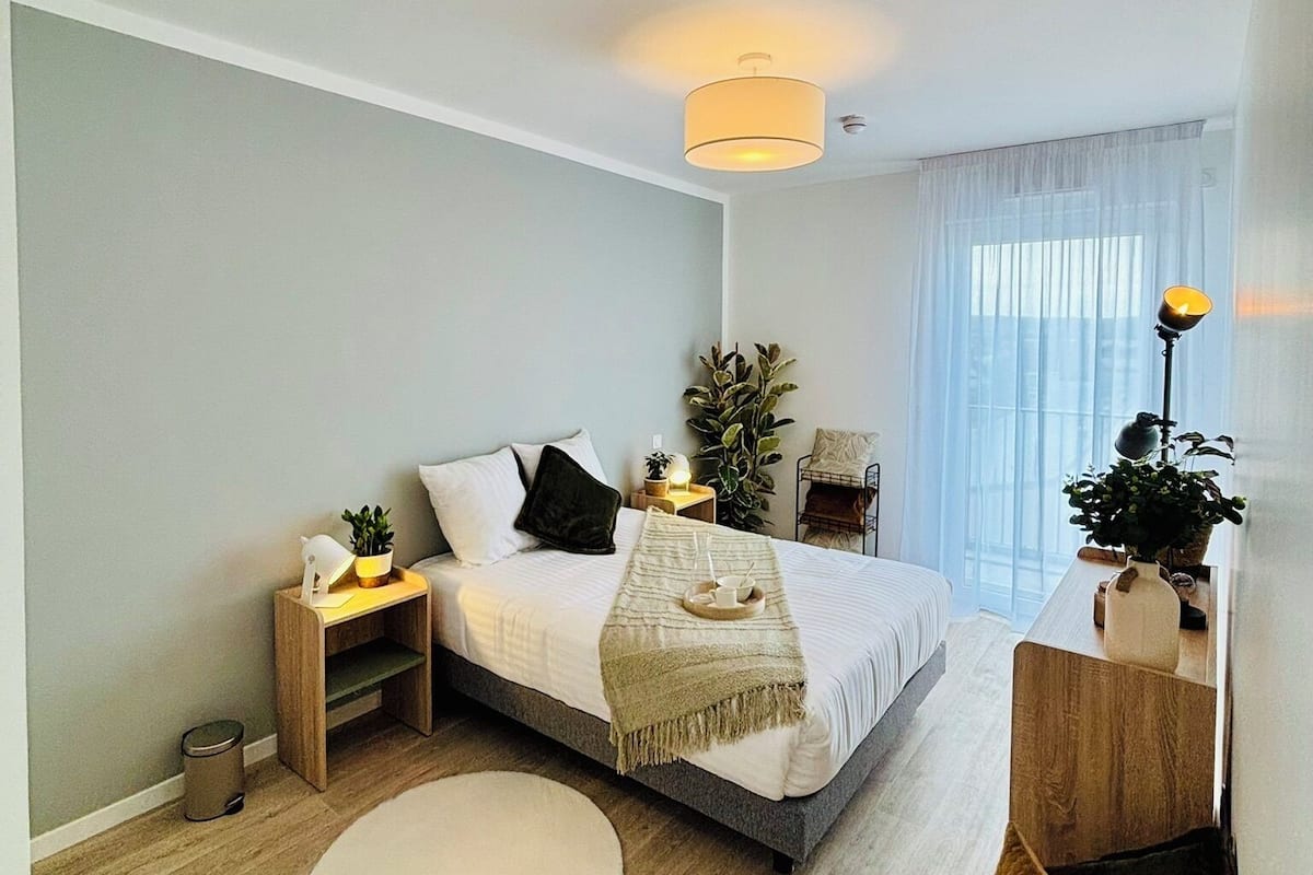 Een moderne slaapkamer met een bed, nachtkastjes, een bedlampje, een potplant en een groot raam met dunne gordijnen.