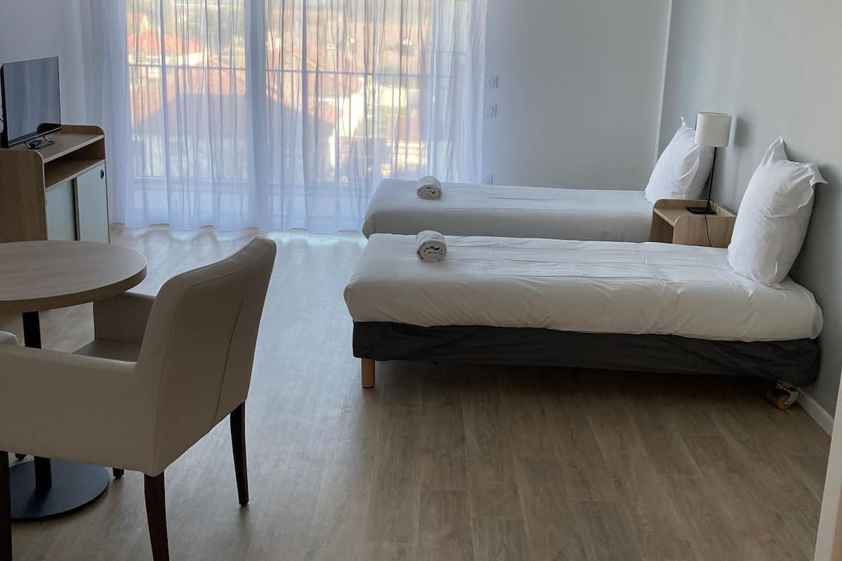 Een hotelkamer met twee bedden, een bureau, een stoel en een groot raam met dunne gordijnen.