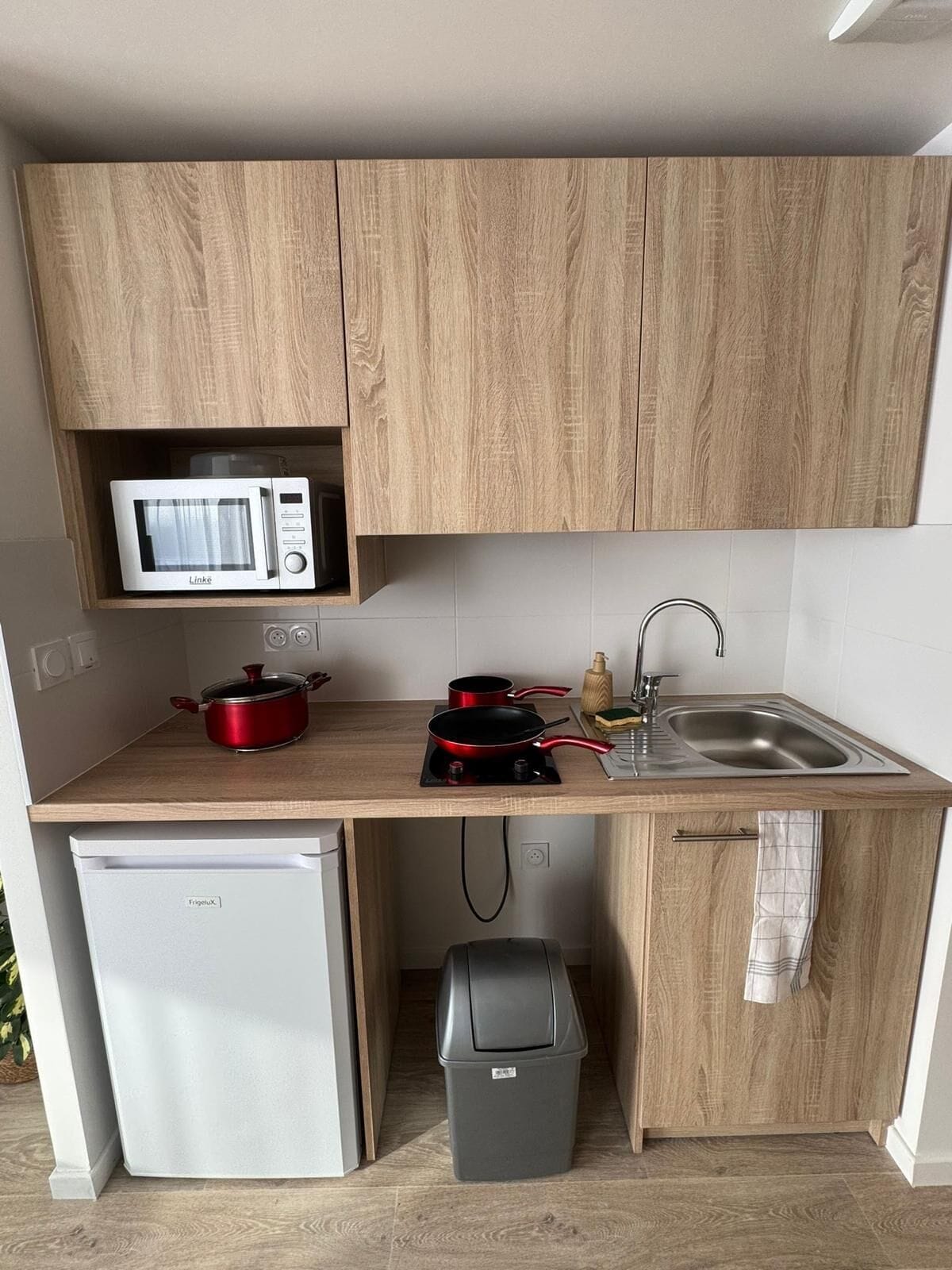 A compact kitchen with a microwave, a small fridge, a sink, and a trash bin.