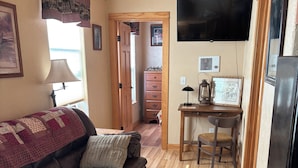 Un salon chaleureux avec un canapé en cuir, un plancher en bois, un téléviseur fixé au mur et un coin cuisine équipé d’un réfrigérateur et d’une table.
