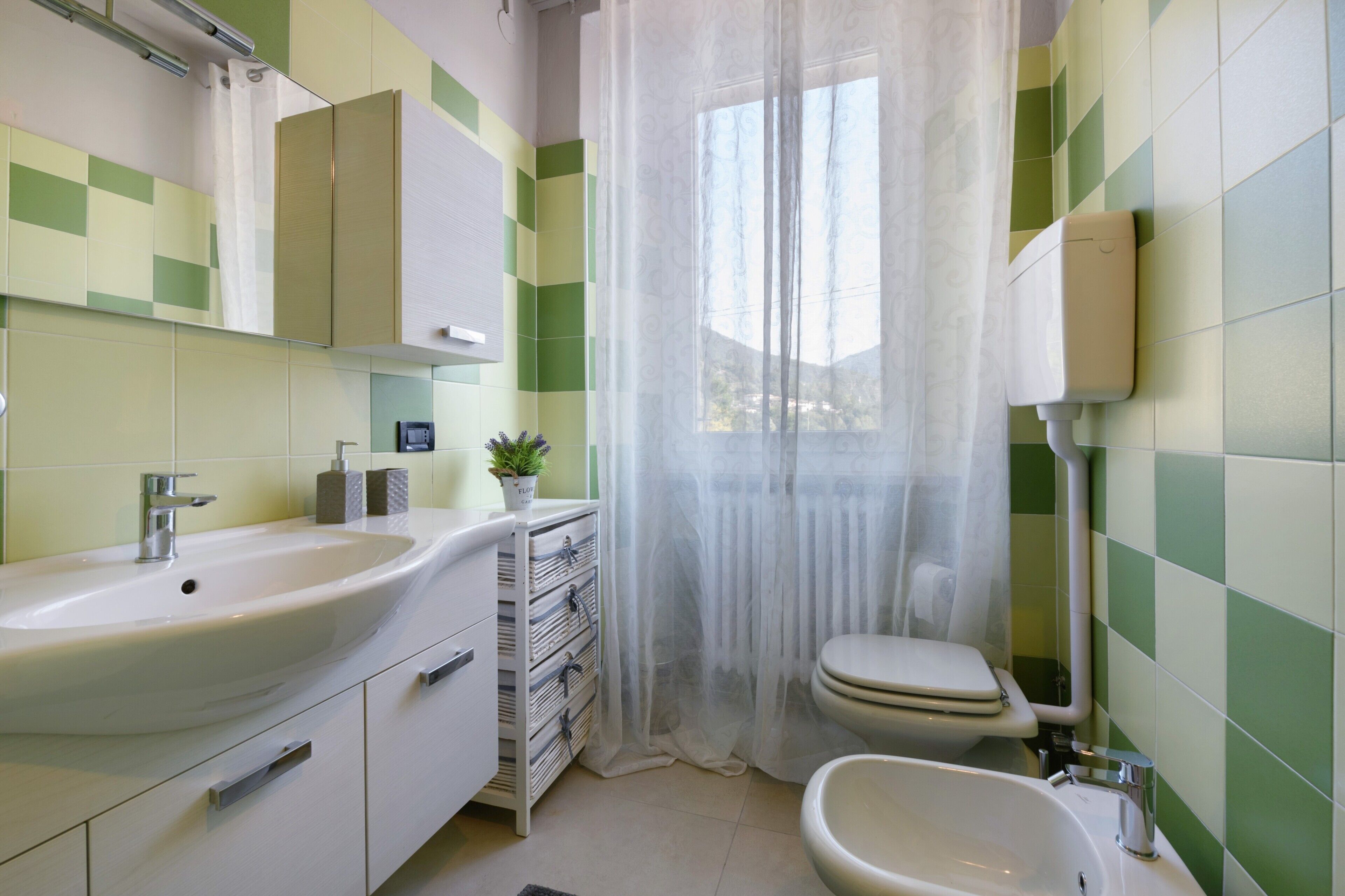 A bathroom with a sink, toilet, bidet, and a window with a sheer curtain.