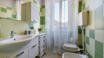 A bathroom with a sink, toilet, bidet, and a window with a sheer curtain.