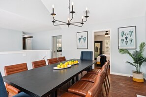 A dining area with a long table, brown leather chairs, and a chandelier. There are framed artworks on the walls and a potted plant on the floor.