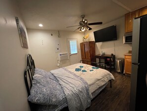 Ein Schlafzimmer mit einem Bett, Deckenventilator, Fernseher, Schreibtisch und einer Küchenzeile mit Kühlschrank.