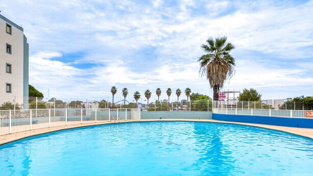 A swimming pool with a white fence, palm trees, and a building in the background.