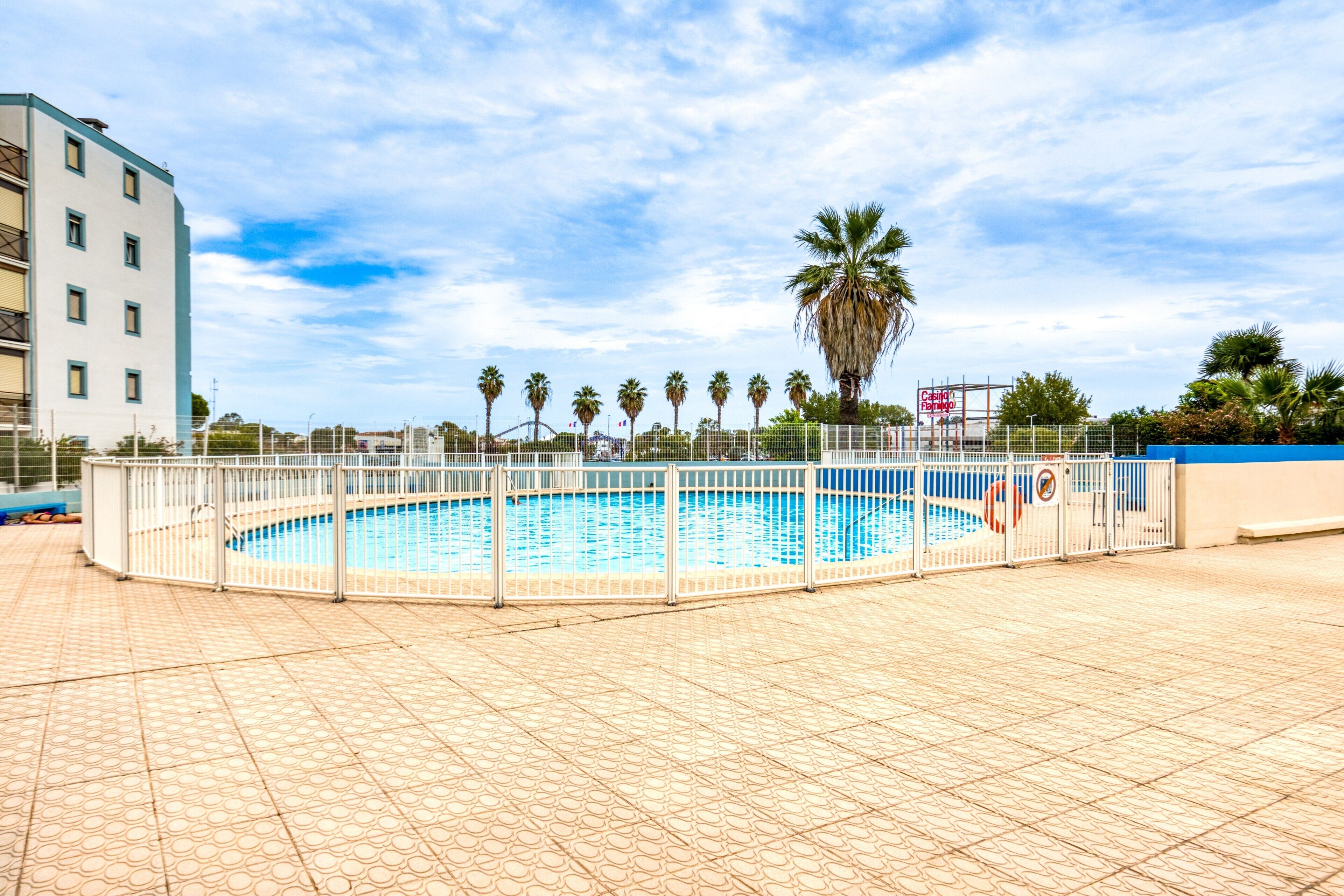 A swimming pool with a white fence, palm trees, and a building in the background.