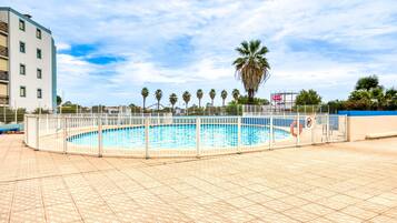 A swimming pool with a white fence, palm trees, and a building in the background.