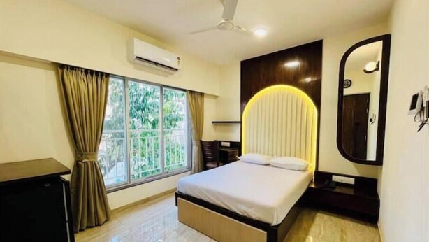 A modern hotel room with a bed, a desk, an air conditioning unit, a window with curtains, a ceiling fan, and a large mirror.