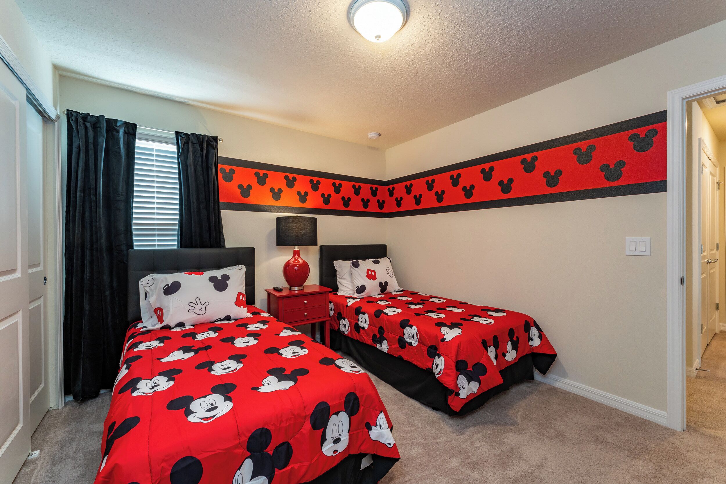 Une chambre avec deux lits, chacun agrémenté d’une couvre-lit aux motifs de Mickey Mouse, une lampe rouge sur une table de chevet et une décoration murale à l’effigie de Mickey Mouse.