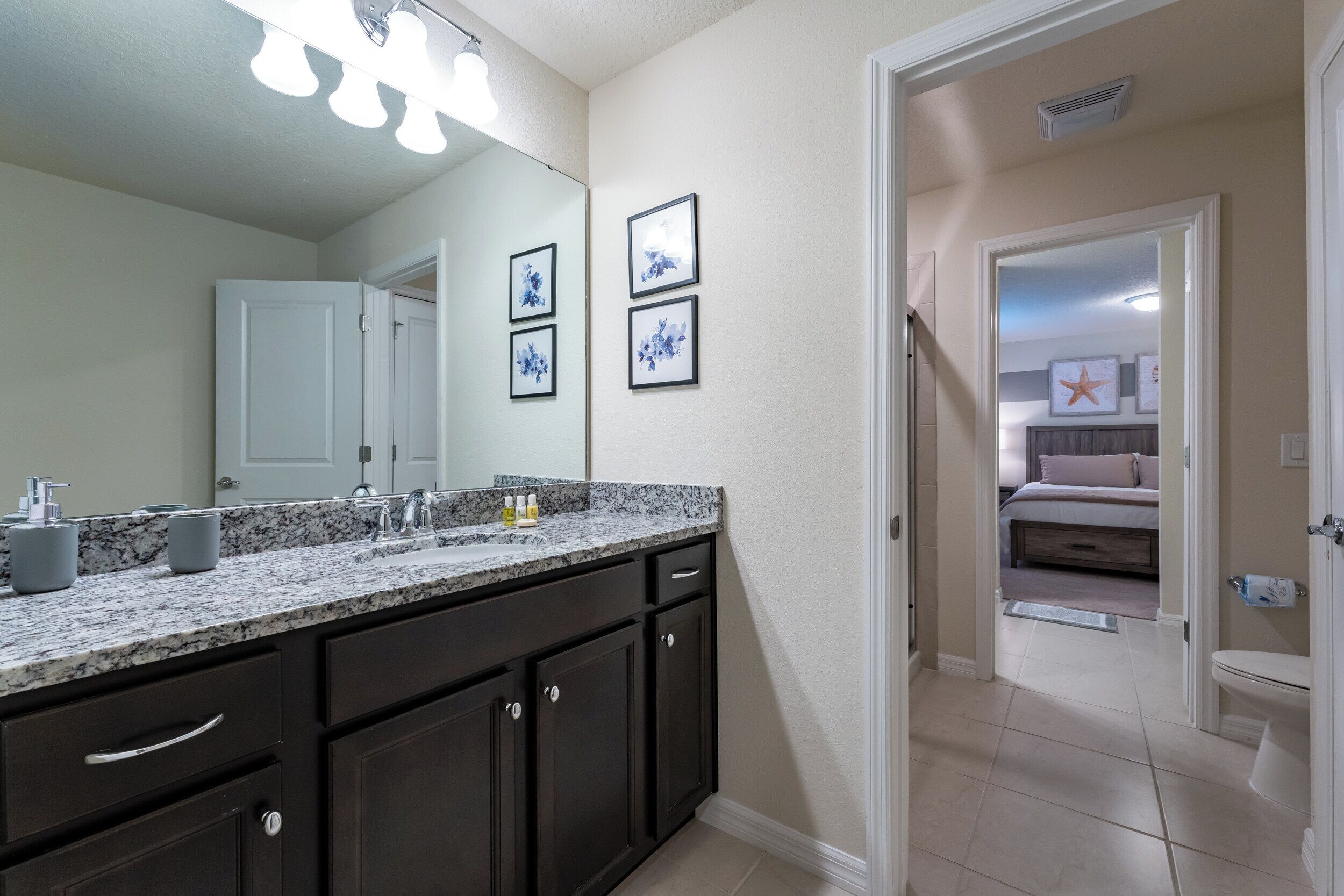 A bathroom with dark cabinets, a granite countertop, and a large mirror. There are framed pictures on the wall and a door leading to a bedroom.