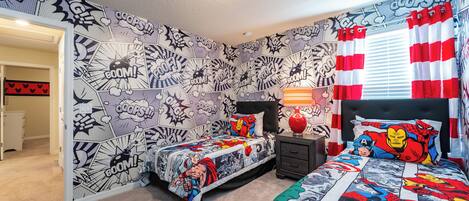 Ein Schlafzimmer mit Comic-Motivelementen an Bettwäsche und Wänden, einem Nachttisch mit Lampe und einem Fenster mit roten und weißen Vorhängen.