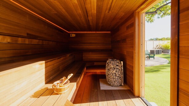 Un sauna aux murs en bois et doté d’un banc, une pile de bois de chauffage et une vue sur une pelouse verdoyante.
