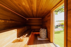 Una sauna con paredes de madera y un banco, leña apilada y vista a un césped verde.