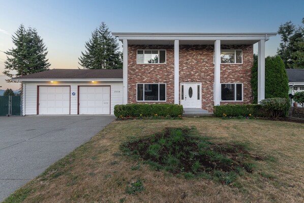 A two-story house with a brick facade, a front garage, and a well-maintained lawn. - Rent-A-Vibe (Abbotsford)