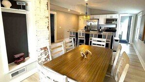 A dining area with a wooden table and white chairs, a kitchen in the background, and a brick wall. - Beautiful Lakehouse close to everything! (Huron)