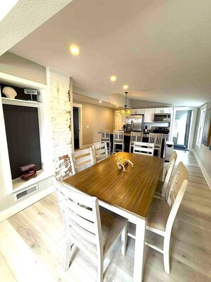Uma sala de jantar com mesa de madeira e cadeiras brancas, cozinha ao fundo e parede de tijolos.
