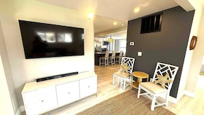A modern living room with a flat-screen TV, white furniture, a dining area in the background, and a dark accent wall. - Beautiful Lakehouse close to everything! (Huron)