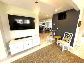 Uma sala de estar moderna com televisão de tela plana, móveis brancos, uma área de refeições ao fundo e uma parede de destaque escura.