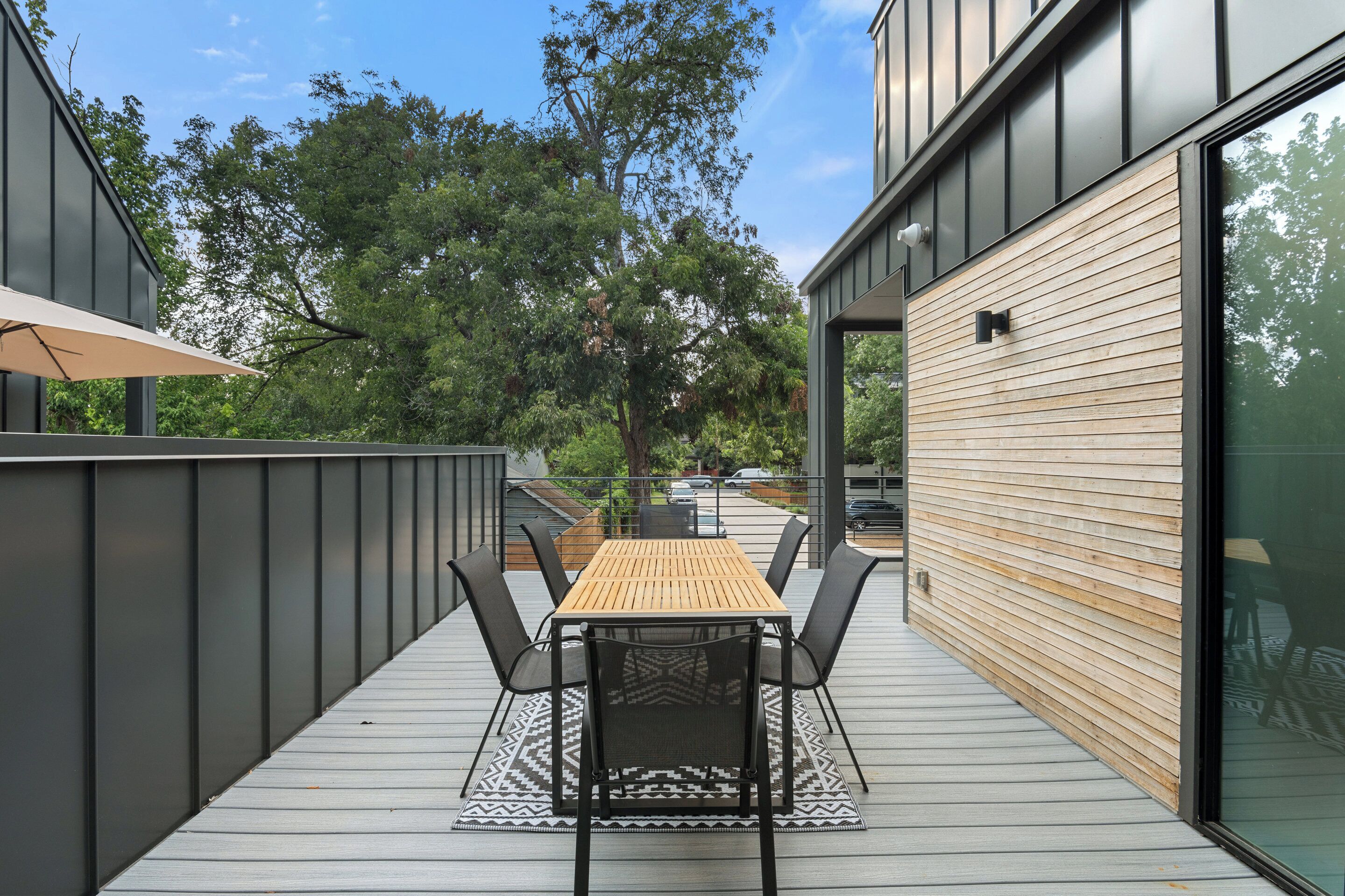 Um deck externo com mesa e cadeiras de madeira, cercado por um corrimão de metal e um painel de vidro.