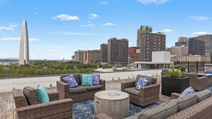 A rooftop lounge with wicker furniture, a round table, and a city skyline view.