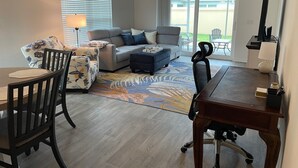 Een woonkamer met een bank, fauteuils, een salontafel en een eettafel met stoelen. Er is een groot raam met zonwering, een plafondventilator en uitzicht op het buiten.