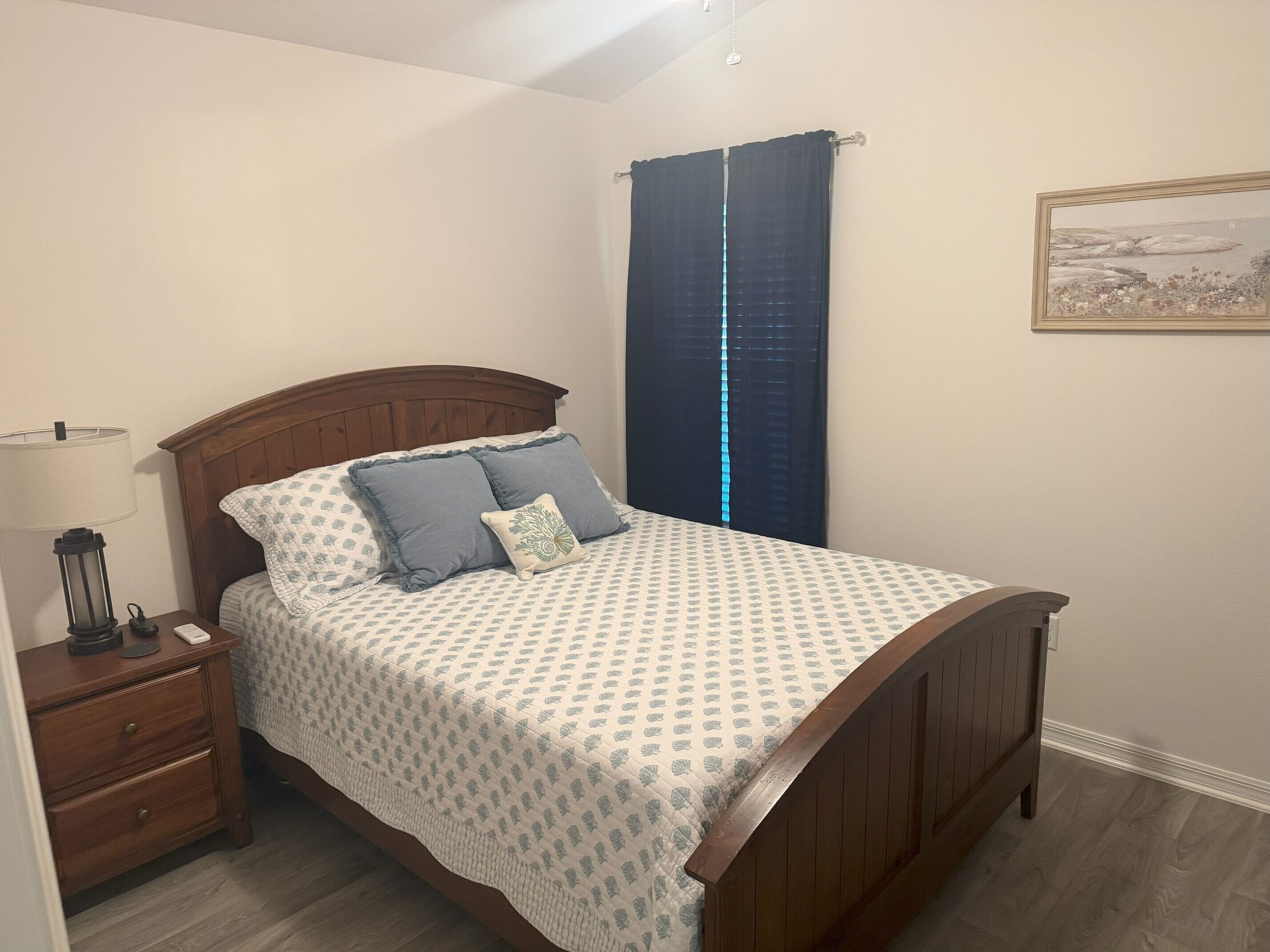 A neatly made bed with a patterned bedspread, a wooden headboard, and two pillows. There is a nightstand with a lamp and a small table beside the bed. A large window with dark curtains is visible in the background.
