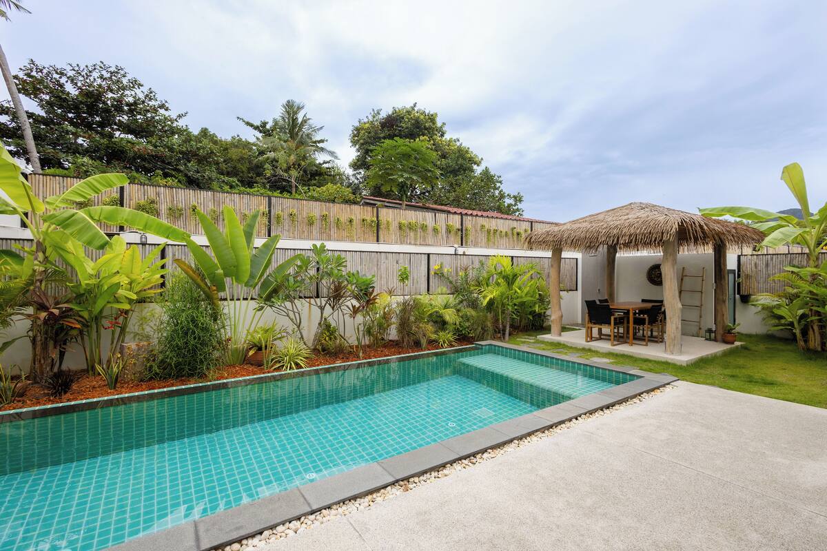 Une piscine dont le bord est revêtu de carreaux, entourée d’une terrasse en béton, d’une clôture en bois et d’une végétation luxuriante, notamment des bananiers.