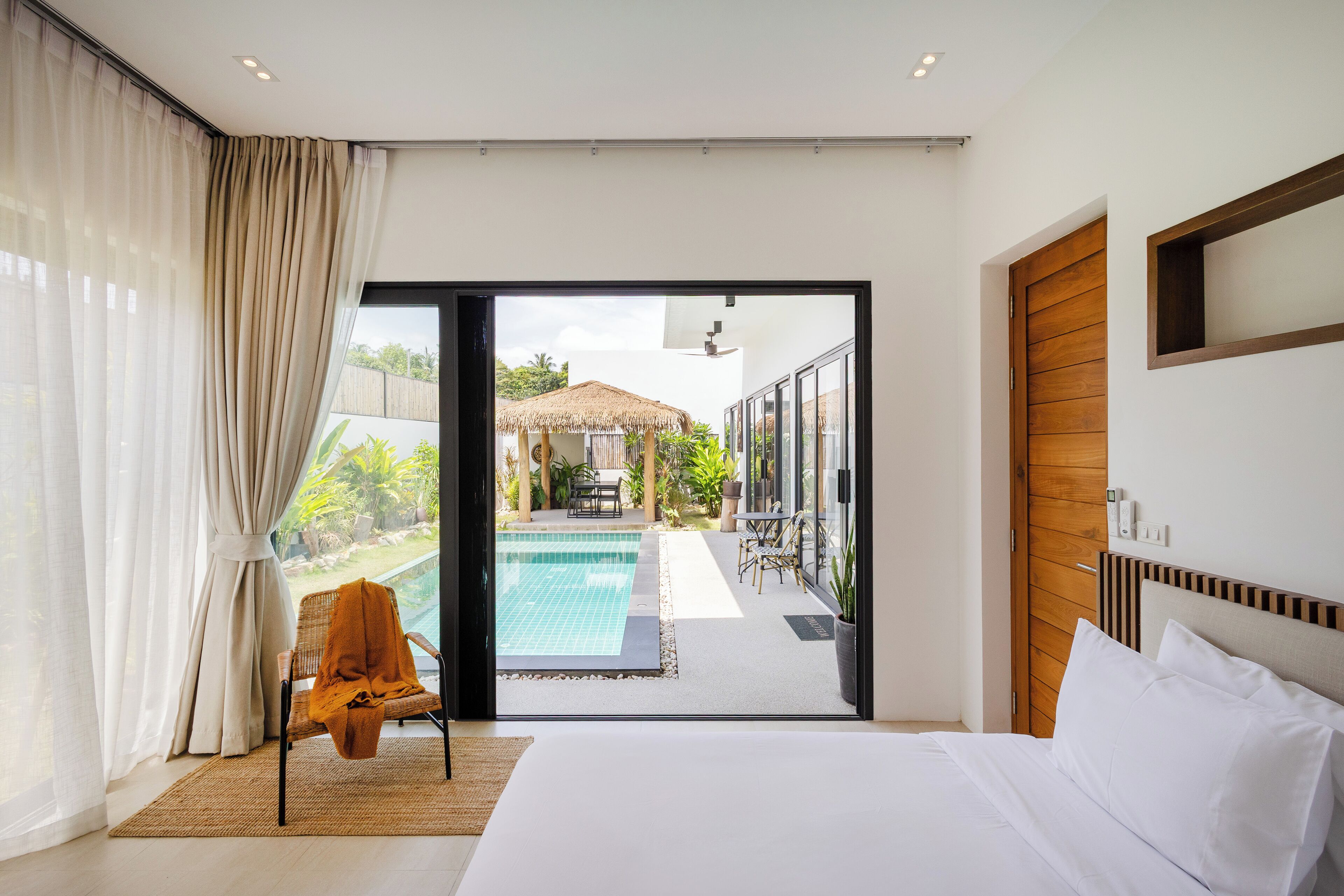 A modern hotel room with a large bed, a view of a swimming pool, and a sliding glass door leading to an outdoor area with a wooden deck and a thatched-roof structure.