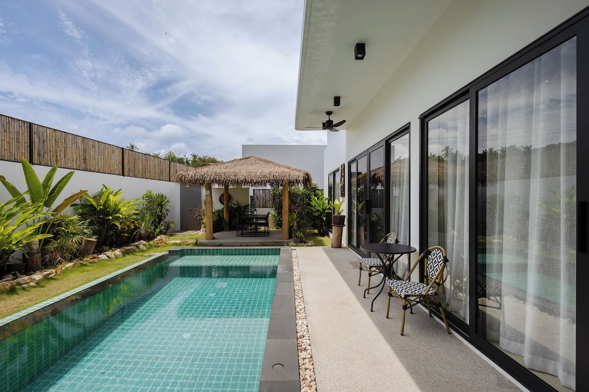 Un espace piscine extérieur moderne, doté d’un gazebo à toit de chaume, d’une table et de chaises, ainsi que d’une grande porte-fenêtre coulissante.