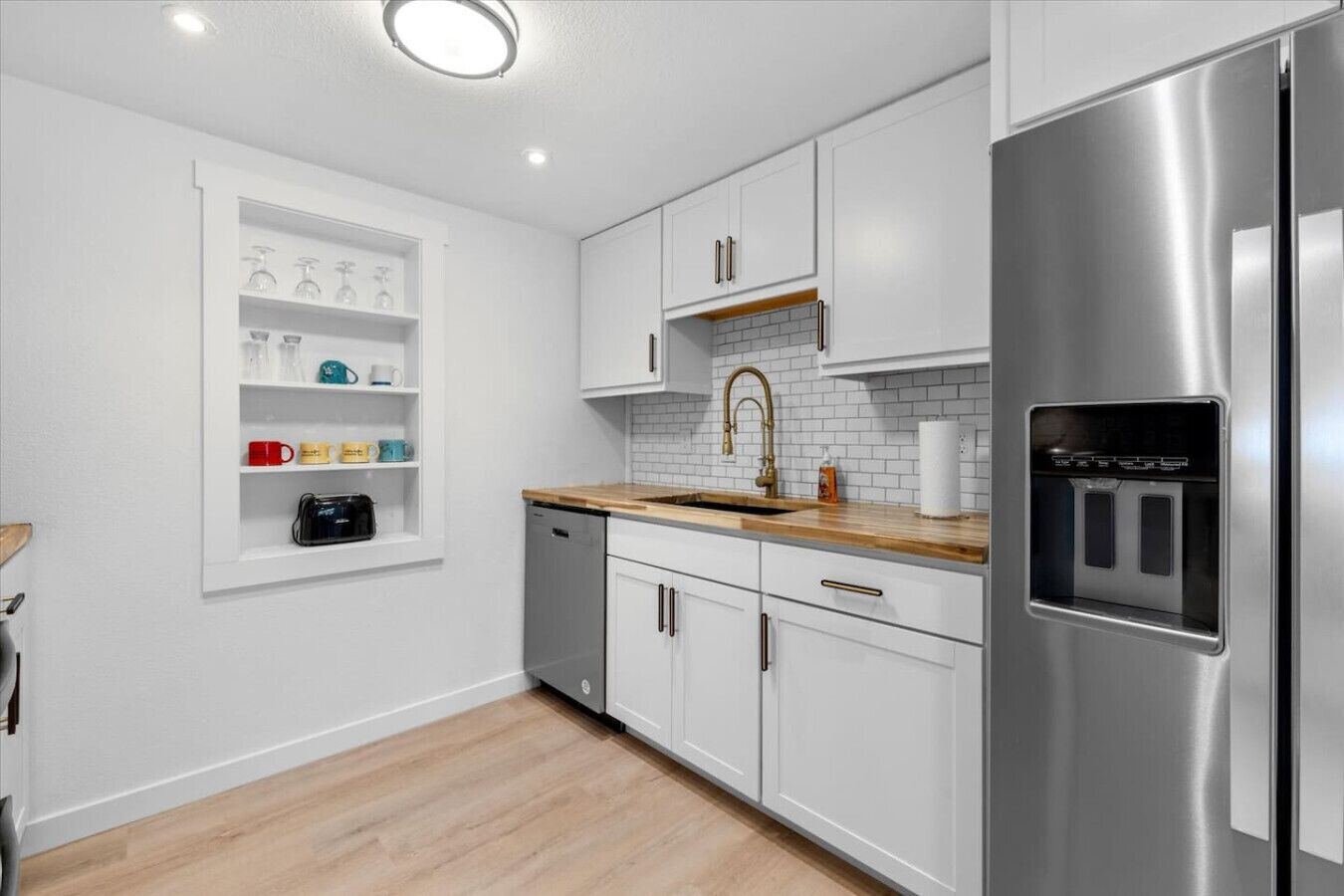 A modern kitchen with white cabinets, a stainless steel refrigerator, a wooden countertop, and a built-in shelf with mugs and a coffee maker.